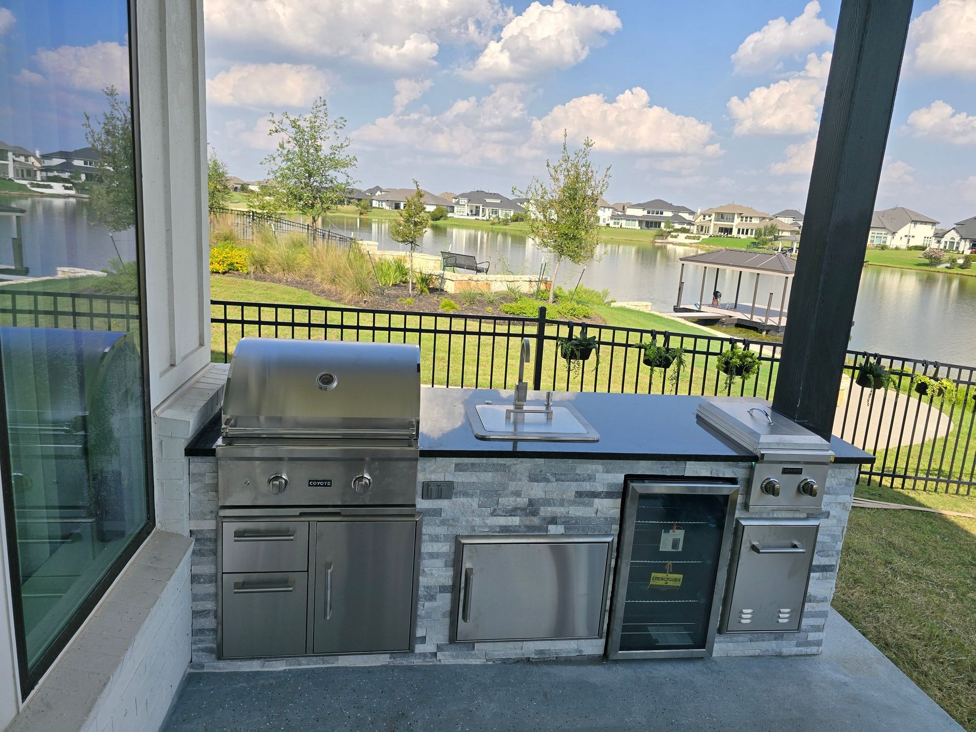 Outdoor kitchen with stainless steel grill and sink overlooking a serene lake and modern homes.